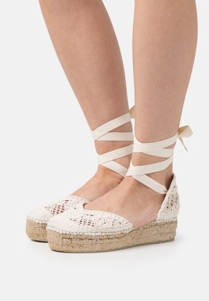Espadrilles de ganchillo blancas con plataforma de yute, que presentan un lazo en los tobillos entrecruzado. Diseño de puntera abierta y tejido texturizado en la parte superior.