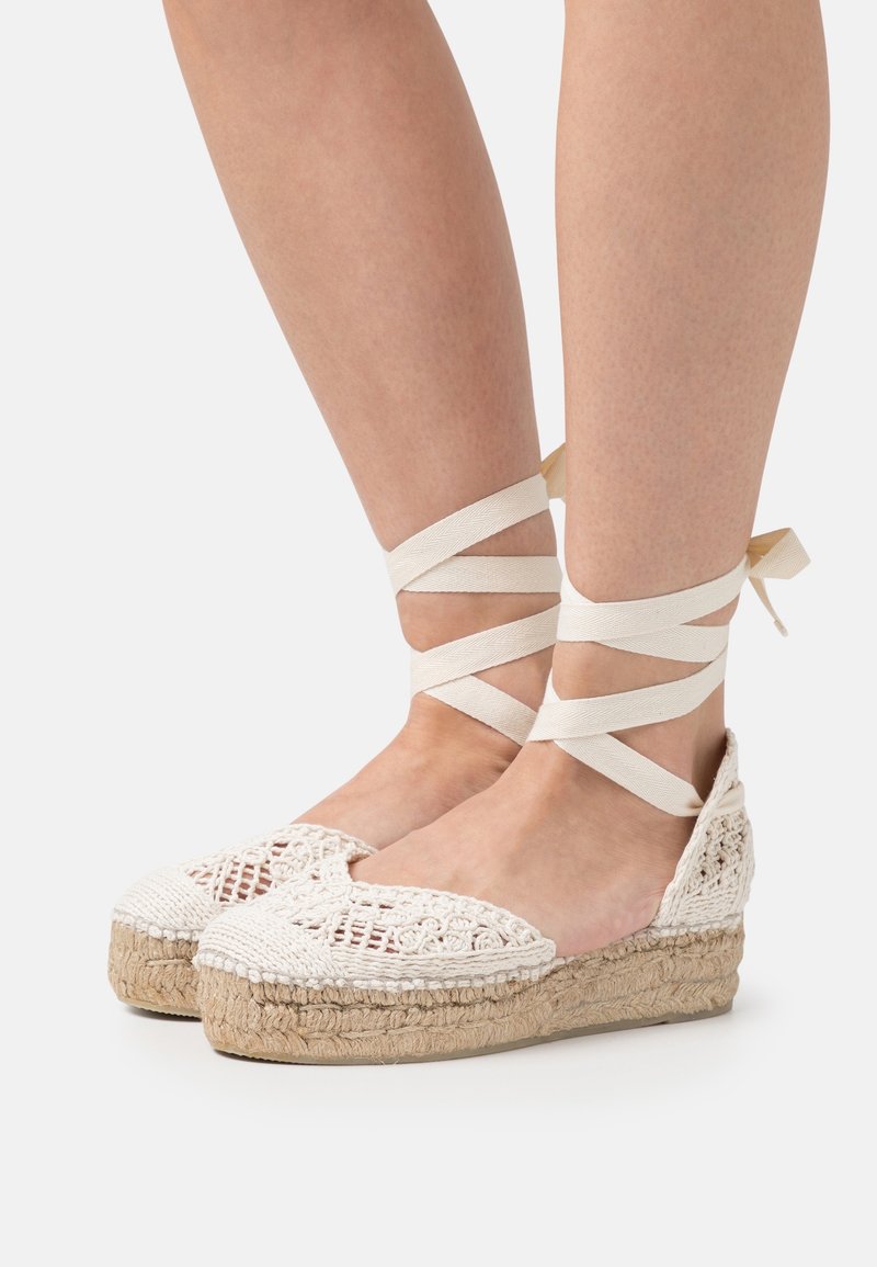 White crochet espadrilles with jute platform, featuring crisscross ankle ties. Open-toe design and textured fabric on the upper.