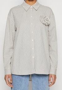 Woman wearing white button-up shirt with thin black vertical stripes and fabric rose detail on left chest, paired with light blue jeans.