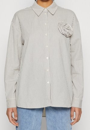 Vrouw in een witte blouse met dunne zwarte verticale strepen en een stoffen rozen detail op de linkerborst, gecombineerd met lichtblauwe jeans.