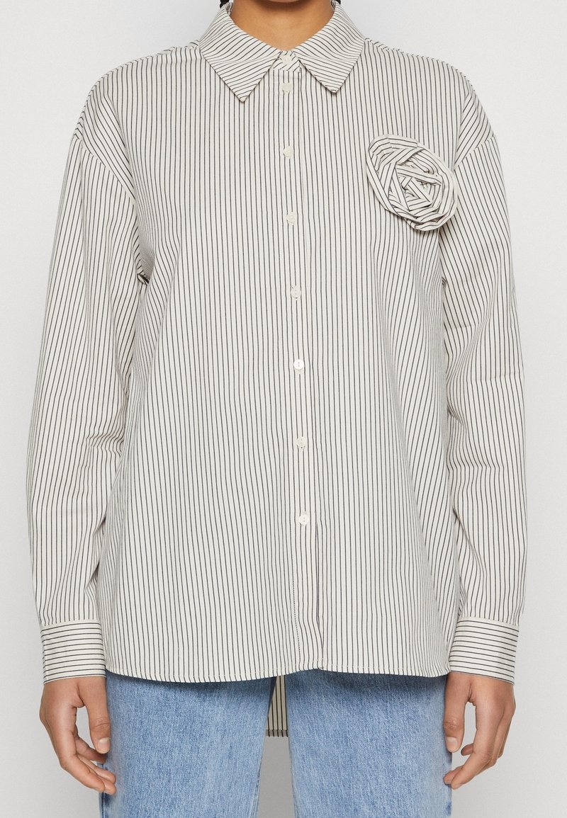 Woman wearing white button-up shirt with thin black vertical stripes and fabric rose detail on left chest, paired with light blue jeans.