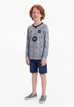 Garçon roux souriant, portant un maillot de football à manches longues gris, un short bleu foncé et des chaussures grises sans lacets, debout sur un fond blanc.
