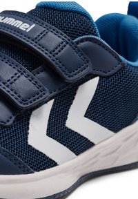 Blå sportskor med meshtyg, två kardborreband och kontrasterande vita zigzag-accenter på sidan. Gummisula med texturerat mönster.