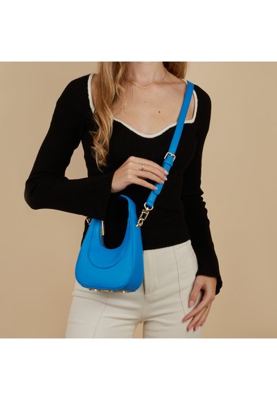 Felblauwe leren handtas met gebogen vorm, bovenste ritssluiting en verstelbare riem; gecombineerd met een zwart geribd topje en crème broek.