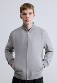 Szürke bomber dzseki sima anyagból, elülső cipzárral, bordázott gallérral és mandzsettákkal, valamint oldalzsebekkel. Egyszerű, modern dizájn.