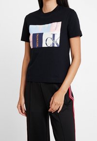Černé bavlněné tričko s boxy střihem, které obsahuje barevnou grafiku s logem Calvin Klein a textem v různých písmových typech.