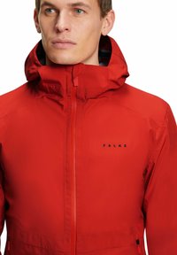 Giacca impermeabile rossa con colletto alto e cappuccio regolabile. Presenta una chiusura con zip e un logo sobrio in nero sul petto.
