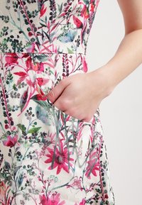 Main reposant dans la poche d'une robe blanche avec un motif floral rose, rouge et vert sur un fond clair.