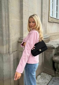 Femme blonde en blazer rose et jean bleu se tient près d'un bâtiment en pierre, portant un sac à main noir matelassé avec une bandoulière chaîne dorée à l'épaule.