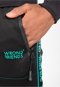 Zwarte joggingbroek met een ritszak. Voorzien van turquoise "WRONG FRIENDS" branding en contrasterende stikdetails. Soepele stoftextuur.