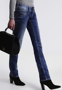 Kvinna i blå jeans, svarta klackstövlar, grå långärmad topp, som håller en svart handväska i sina händer.