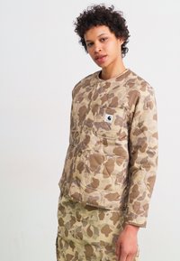 Quilted camouflage jacket in beige and brown tones with a snap closure, long sleeves, and a front pocket featuring a brand logo.