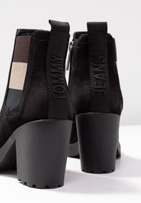 Bottes de cheville en daim noir avec un talon épais. Comprend des panneaux élastiques de teintes brunes et beiges, avec des logos lisant "TOMMY" et "JEANS."