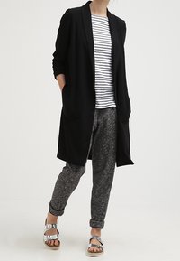 Langer schwarzer Cardigan über einem gestreiften weißen und marineblauen Oberteil, kombiniert mit grauen, strukturierten Hosen und silbernen Sandalen. Schlichte, lässige Outfit.