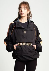 Ung kvinde i en sort og beige OAK25 jakke med hænderne i frontlommen, der kigger til siden mod en enkel lys baggrund.