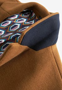Col du manteau en laine marron avec un accent bleu marine, montrant une doublure intérieure à motifs géométriques colorés.