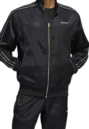 Czarna satynowa kurtka sportowa z wysokim kołnierzem, złotym zamkiem błyskawicznym oraz trzema białymi paskami wzdłuż każdego rękawa. Złote logo Adidas na klatce piersiowej.