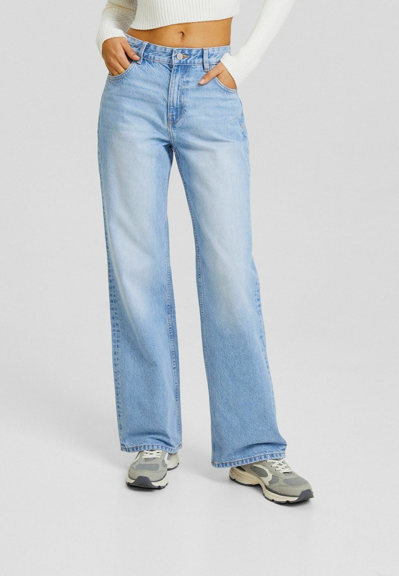 Personne portant un jean large bleu clair avec les mains dans les poches et des baskets blanches, le haut à manches longues blanc court est visible.
