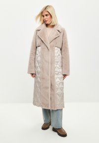 Kvinna med blont hår som bär en lång beige kappa med quiltade sidor, ljusblå jeans och bruna stövlar, stående mot en vit bakgrund.