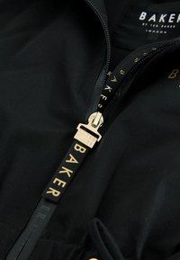 Čierny zips na bunde so zlatou textilnou pútkom „BAKER“ a zlatým kovovým posuvníkom s vyrytým nápisom „TED BAKER“, viditeľná značková vnútorná etiketa.