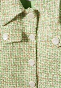 Giacca in tweed verde, beige e bianco con colletto a punta, tasche a patta e sei grandi bottoni rotondi color crema sulla parte anteriore.