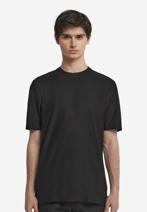 Junger Mann mit kurzen braunen Haaren, der ein schlichtes schwarzes T-Shirt mit kurzen Ärmeln und Rundhalsausschnitt trägt, steht vor einem weißen Hintergrund.