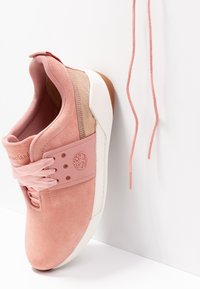 Baskets en suède rose avec semelle blanche et bride en caoutchouc, un lacet partiellement défait et pendant contre un fond blanc.