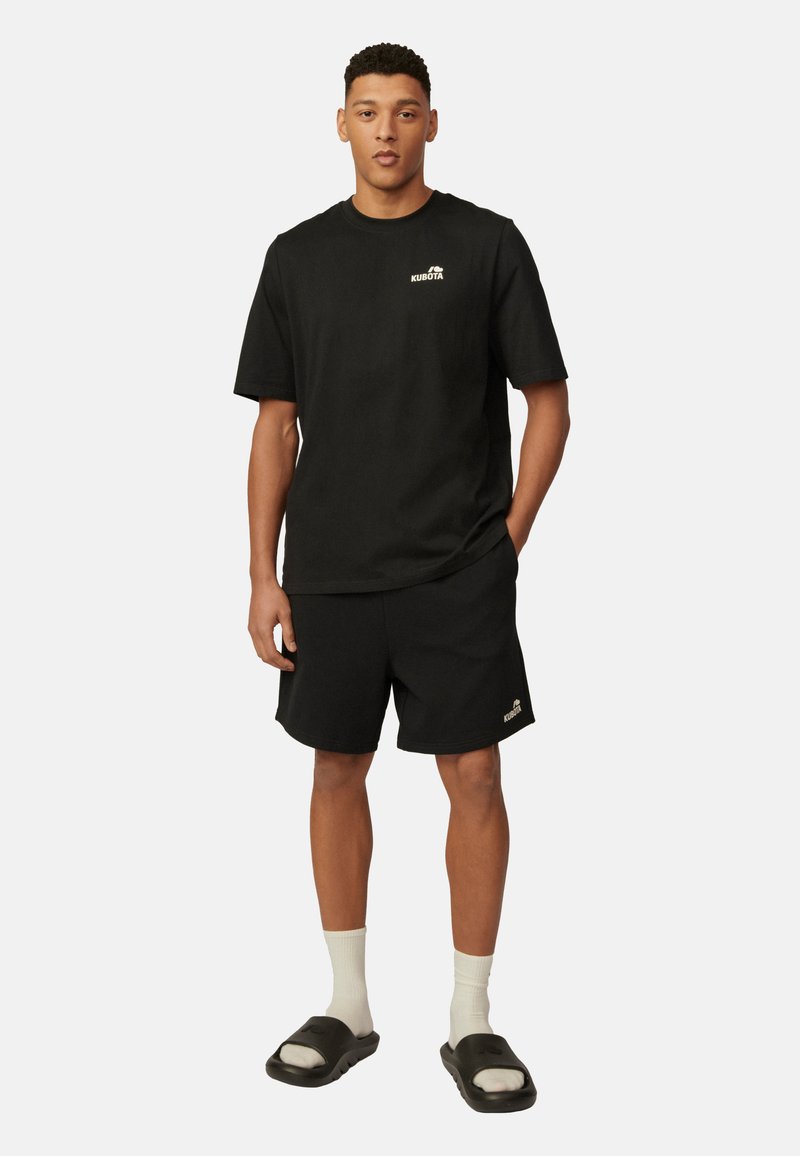Czarna bawełniana koszulka z logo na piersi, w połączeniu z czarnymi szortami i sandałami wsuwanymi, uzupełniona białymi skarpetkami do kostek.