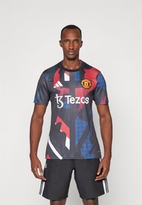 Črno-siva nogometna majica z drznimi rdečimi in modrimi abstraktnimi vzorci, ki na sprednji strani vsebuje logo Manchester United in "Tezos".