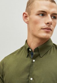 Olive zöld gombos ing gallérral és fehér gombokkal. A gallér belsejében mintás szövet részlet található. Simára szövött anyag.