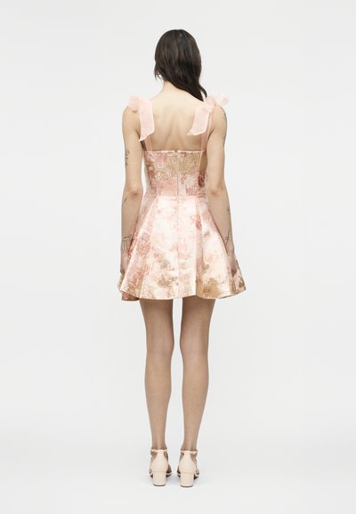 Femeie purtând o rochie roz florală scurtă, fără mâneci, cu bretele în formă de fundă, cu faţa spre spate, purtând pantofi bej cu toc gros, pe un fundal alb.