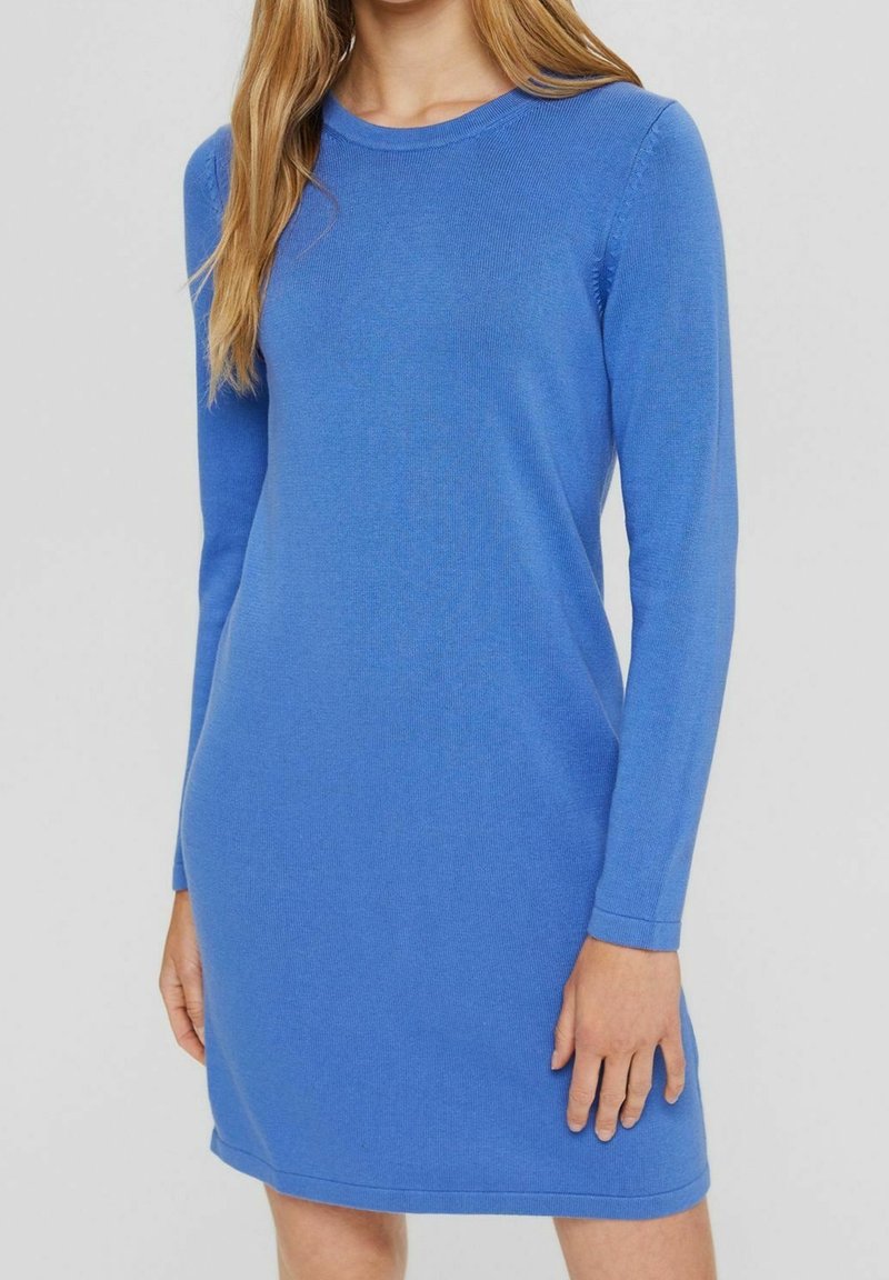 Femme portant une robe en maille bleue cintrée, manches longues, longueur genou, avec un col rond, sur un fond gris uni.