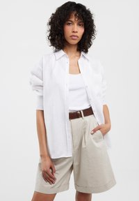 Camisa blanca de lino de manga larga sobre una camiseta blanca sin mangas, combinada con pantalones cortos de tela con pliegues en color beige y un cinturón marrón. La camisa tiene detalles de botones.