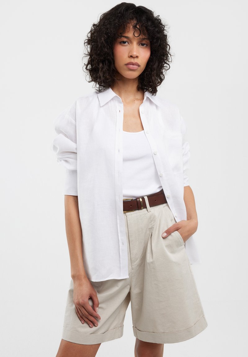 Camisa blanca de lino de manga larga sobre una camiseta blanca sin mangas, combinada con pantalones cortos de tela con pliegues en color beige y un cinturón marrón. La camisa tiene detalles de botones.