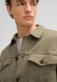 Giacca in canvas khaki con colletto a punta, tasche frontali con pattina e chiusura a bottoni. Tessuto semplice con dettagli di cucitura visibili.