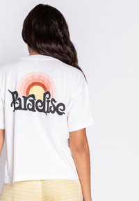 Bílé zkrácené tričko s nápisem "Paradise" v tučném černém písmu, s gradientním duhovým designem v jemných odstínech růžové, oranžové a žluté na zadní straně.