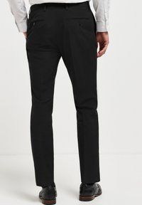 Schwarze formelle Hosen mit taillierter Passform, ausgestattet mit Gesäßtaschen, Knopfverschlüssen und einer glatten Textur. Tragen mit schwarzen Schnürschuhen.