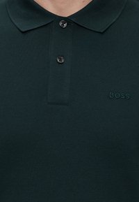 Mørkegrønn poloskjorte med to svarte knapper festet, krage og "BOSS" brodert på venstre bryst.