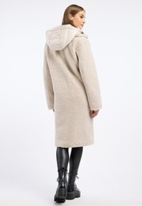 Crème-kleurige jas met een pluche textuur, voorzien van een gewatteerde capuchon, lange mouwen en een rechte silhoët. Draagt over zwarte leggings en laarzen.