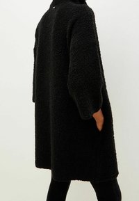 Cappotto oversize nero realizzato in tessuto testurizzato e morbido; presenta un colletto alto e maniche larghe. Tasche laterali sono visibili.