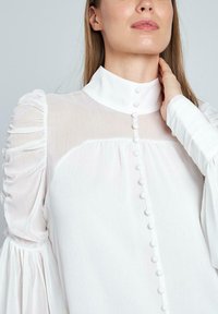 Blusa blanca con cuello alto, tejido transparente, mangas abullonadas y detalles de botones. Presenta un material suave y texturizado, con sutiles pliegues.