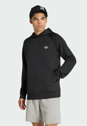 Młody mężczyzna w czarnej bluzie z kapturem z logo Adidas, jasnoszortach i czarnej czapce z daszkiem, stoi z jedną ręką w kieszeni.