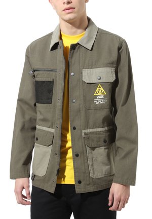 Jeune homme portant une veste Vans verte et beige avec plusieurs poches avant sur une chemise jaune, montré des épaules aux hanches.