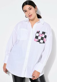 Camicia bianca con maniche lunghe e taschino a sinistra, caratterizzata da un motivo a scacchi bianco e nero con cuori rosa.