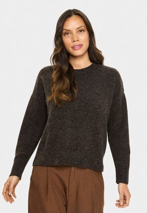Femme aux longs cheveux ondulés portant un pull en tricot marron foncé et un pantalon couleur rouille, se tenant contre un fond clair uni.