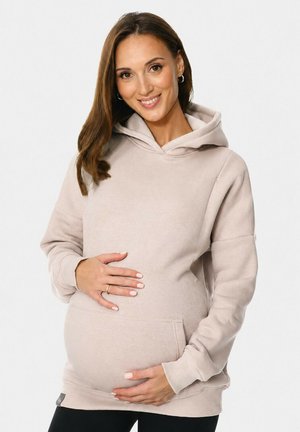 Lächelnde schwangere Frau in einem beigefarbenen Hoodie wiegt ihren Babybauch sanft mit beiden Händen vor einem schlichten hellen Hintergrund.