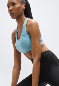 Γυναίκα με αθλητικό σουτιέν Puma σε ανοιχτό γαλάζιο και μαύρο κολάν, τεντώνεται με τα χέρια στους μηρούς μπροστά σε απλό ανοιχτό φόντο.