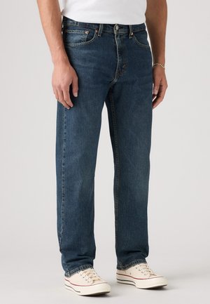 Homme portant un jean bleu droit, des baskets blanches basses et une chemise blanche, debout devant un fond uni.