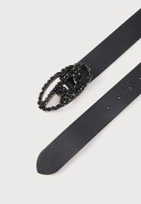 Melns ādas jostas ar dekoratīvu ovālu sprādzi, uz kuras ir melnu dārgakmeņu klasteris pinuma dizainā. Gluda tekstūra, ar nokalpotu galu.