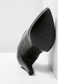 Bota de tacón de cuero negro con punta afilada apoyada contra una pared blanca, con la suela y el tacón claramente visibles y un patrón de textura en la suela.